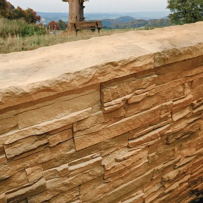 Revestimiento de piedra Petra Mineral - Cantidad total: 4 pzs., Mineral - Primer plano de una pared de piedra decorativa con un banco y árboles al fondo, ofreciendo una estética rústica y natural.