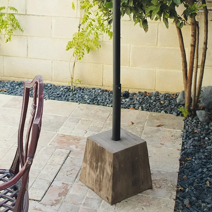 Base de parasol cuadrada - L x An: 26 x 18 cm - Base de sombrilla de hormigón con un poste negro, situada en una zona ajardinada con plantas y rocas.