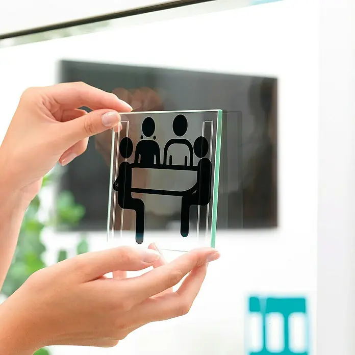 Tesa Powerbond Cinta de doble cara Transparente - 19 mm x 5 m - La imagen muestra una señal transparente con un pictograma negro de una reunión, sostenida por manos contra un fondo borroso.