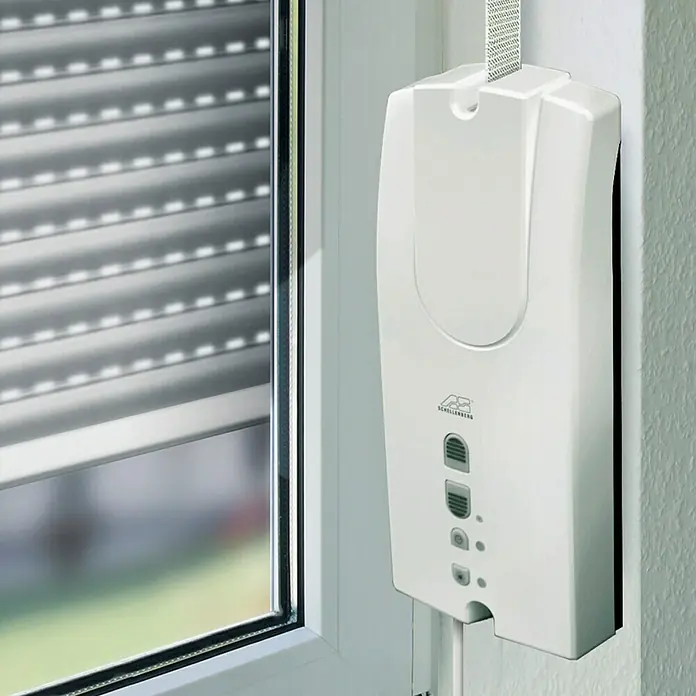 Schellenberg Recogedor de cinta de persiana Rollodrive 55 - En pared, Anchura de la correa: 14 mm - 23 mm, 230 mm - Primer plano de un accionamiento de persiana enrollable Schellenberg para ventanas, montado en una pared blanca y un marco de ventana.