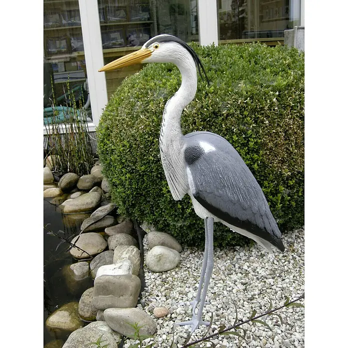 Ubbink Tiervertreiber Fischreiher - 21,5 x 54 x 87 cm, Kunststoff - Die Fotografie zeigt eine dekorative Reiherfigur aus Kunststoff in einem Garten neben einem kleinen Teich und einem grünen Busch.