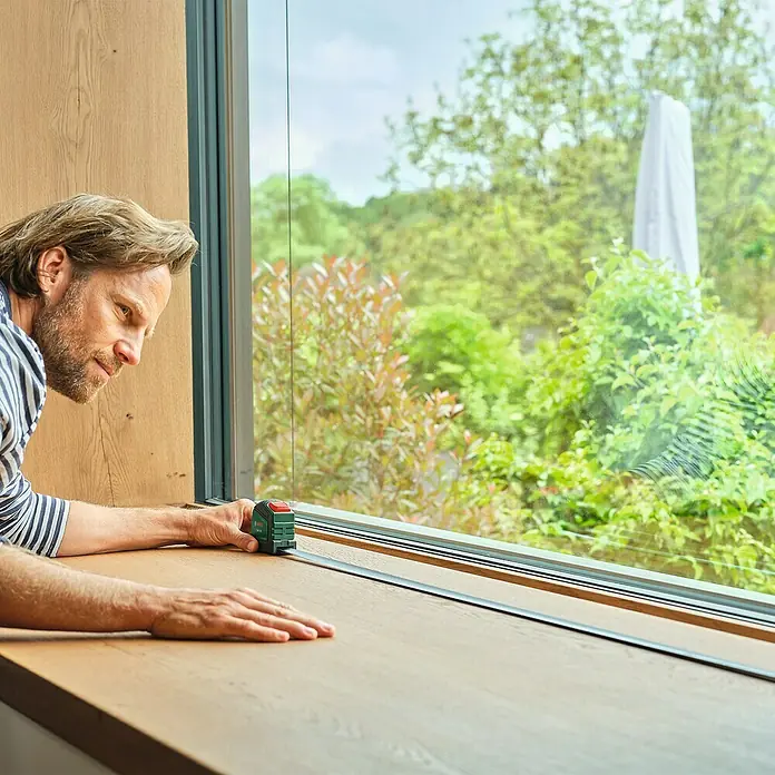 Bosch Bandmaß - 2 m - Mann misst Fensterbank mit einem grünen Bosch-Laser-Entfernungsmesser für präzise Ergebnisse. Ideal für Renovierungsarbeiten.