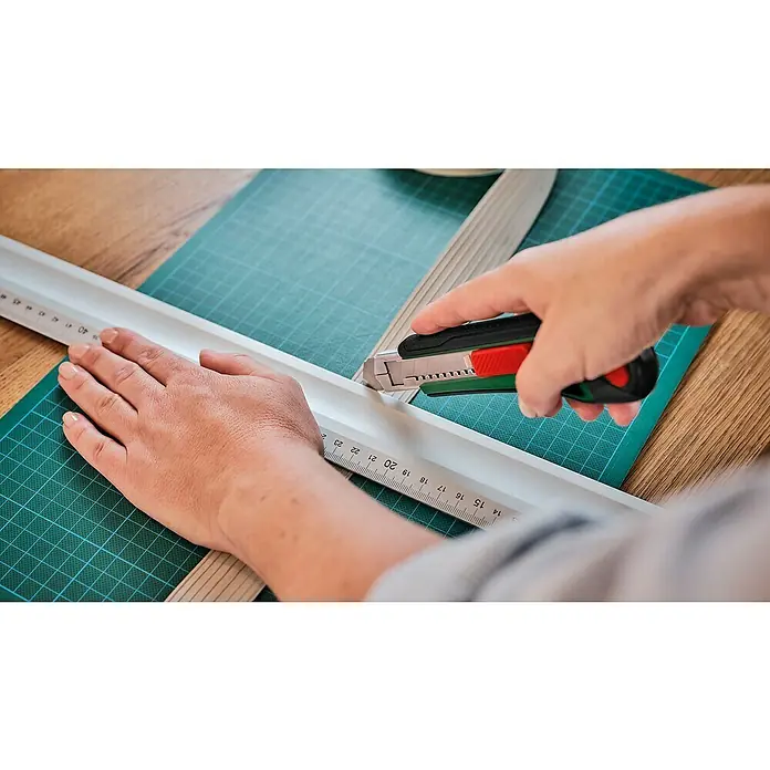 Bosch Abbrechklinge - Passend für: Bosch Cuttermesser, 10 Stk. - Nahaufnahme von Händen, die mit einem Cutter und einem Lineal ein Material auf einer grünen Schneidematte schneiden.