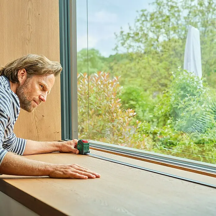 Bosch Bandmaß - 5 m - Ein Mann misst mit einem grünen Rollbandmaß ein Fensterbrett aus Holz. Das Werkzeug ist gut sichtbar.