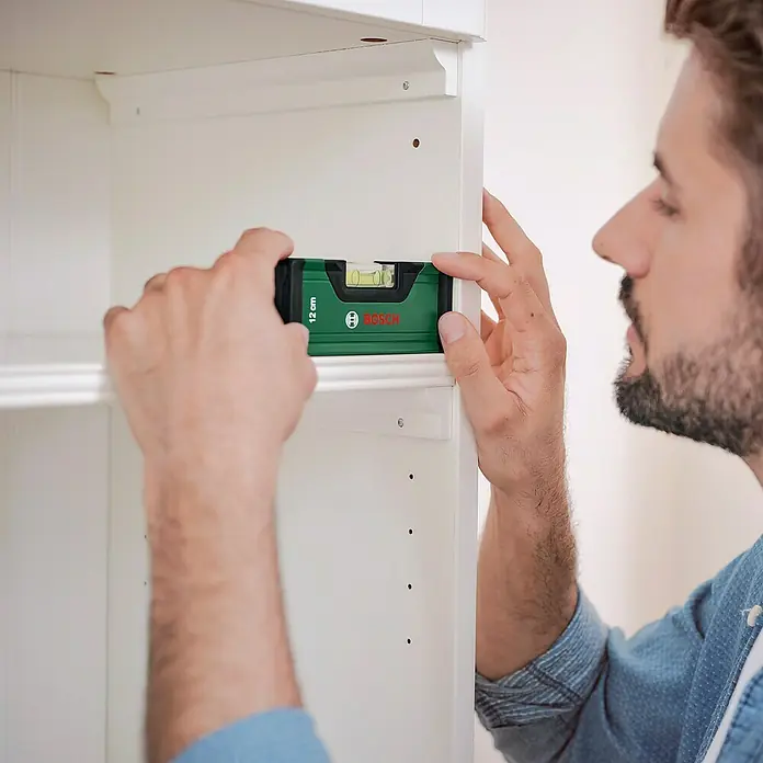 Bosch Wasserwaage - Länge: 12 cm, Anzahl Libellen: 1 Stk. - Ein Mann benutzt eine grüne Bosch Wasserwaage, um ein Regal in einem weißen Schrank auszurichten. Praktisches Werkzeug für Heimwerker.