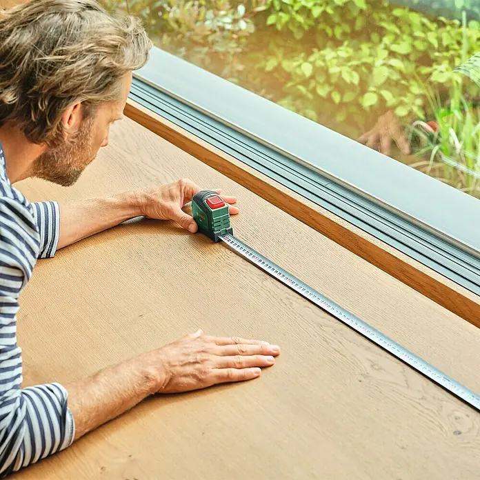 Bosch Bandmaß - 5 m - Ein Mann misst mit einem Bosch-Rollbandmaß eine Oberfläche. Das Bandmaß ist grün und rot und zeigt präzise Messwerte.
