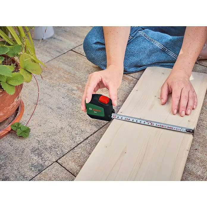 Bosch Bandmaß - 3 m - Nahaufnahme eines Bosch 3m Rollbandmaßes, das ein helles Holzbrett ausmisst, um Präzision und Funktionalität bei DIY-Projekten hervorzuheben.
