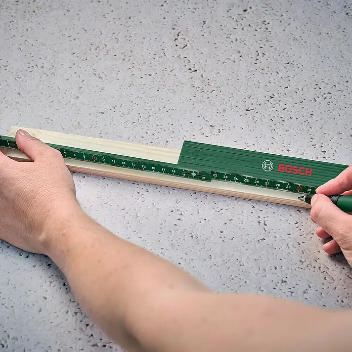 Bosch Zollstock - Länge: 2 m - Eine grüne Bosch-Zollstock liegt auf einem Holzbrett, eine Hand markiert mit einem Stift eine Stelle auf dem Brett entlang des Zollstocks.
