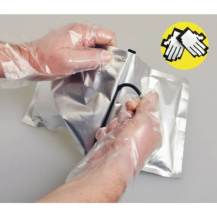 Kaiser Feuchtraum-Abzweigdose Installations-Set Outdoor IP68BOX - 95 x 95 x 55 mm, Aufputz, IP68 - Detailaufnahme einer Person, die eine versiegelte Silberfolienverpackung mit transparenten Handschuhen öffnet.
