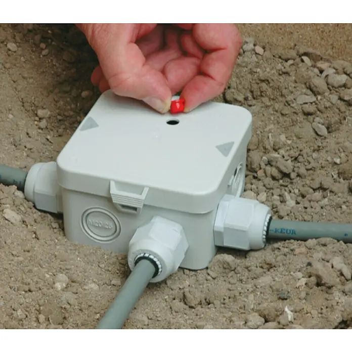 Kaiser Feuchtraum-Abzweigdose Installations-Set Outdoor IP68BOX - 95 x 95 x 55 mm, Aufputz, IP68 - Einbaut IP68 Verteilerdose mit einer Hand, die einen roten Knopf drückt, während sie im Sand eingebettet ist.