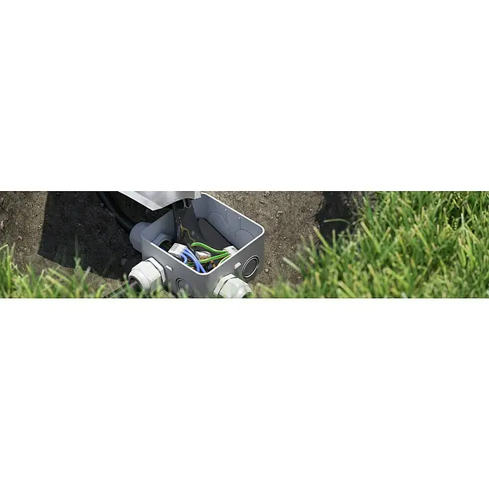 Kaiser Feuchtraum-Abzweigdose Installations-Set Outdoor IP68BOX - 95 x 95 x 55 mm, Aufputz, IP68 - Diese Aufnahme zeigt eine geöffnete graue Abzweigdose mit diversen Kabeln. Sie ist in einen Garten mit Erde und Gras eingebettet.