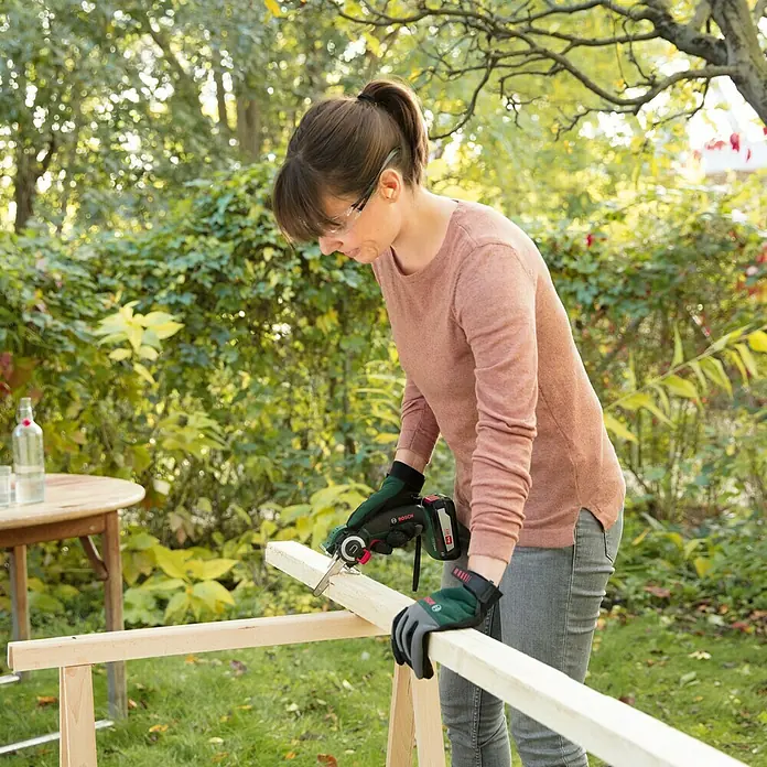 Bosch Power for All 18V NanoBlade-Säge AdvancedCut 18Leerlaufdrehzahl: 7.000 U/min, 18 V, Ohne Akku - Frau arbeitet im Garten mit einer Bosch Akku-Säbelsäge und schneidet ein Holzbrett. Schutzbrille und Handschuhe werden getragen.