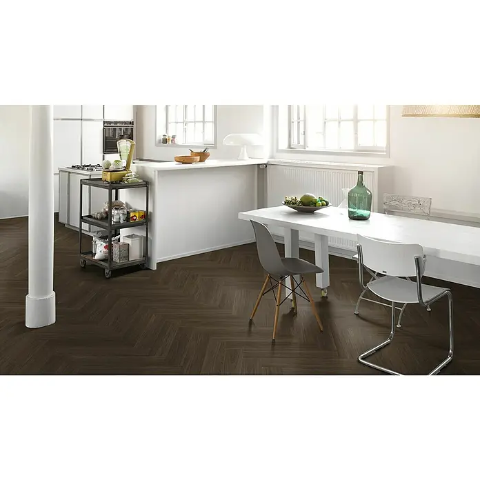 Parador Vinylboden Trendtime 3 - Eiche Oxford dunkelbraun, Fischgrät, 730 x 146 x 5 mm - Ein helles Interieur mit dunklem Fischgrätparkettboden. Eine Küche mit Kochinsel und ein Esstisch mit Stühlen sind zu sehen.