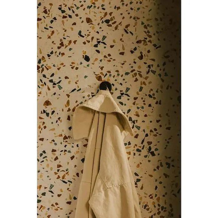 Cosmic Colgador Dual Start - Negro, Latón, Brillante - Chaqueta beige colgada de un gancho oscuro contra una pared de terrazo. La chaqueta es bien visible e invita a comprarla.