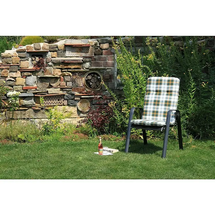 beo Gartenmöbel-Auflage Ejina - Hochlehner, 6 Stk., L x B x H: 114 x 47 x 5 cm, Baumwolle, Polyester, Hellgrün - Gartenszene mit einem bequemen Gartenstuhl mit kariertem Muster, einer Flasche und Büchern auf dem Rasen vor einer kunstvollen Steinmauer.