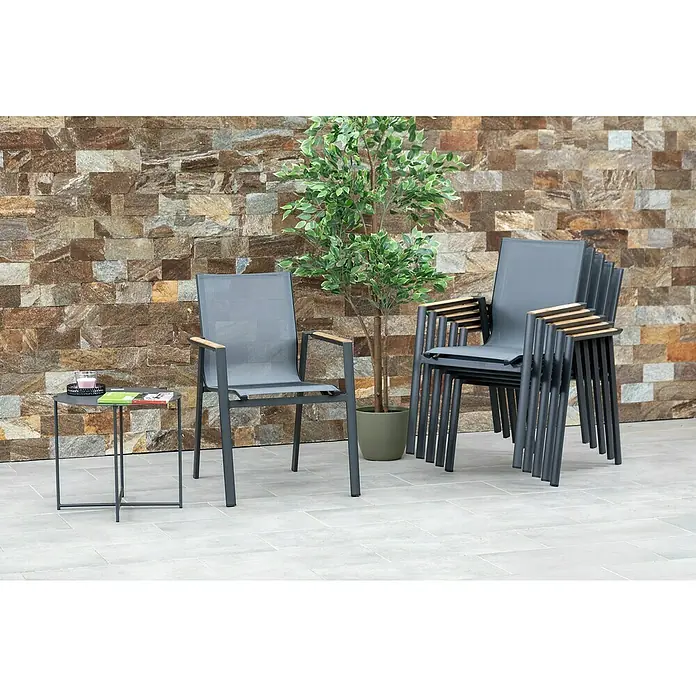 Acamp Stapelsessel Vegas - 4 Stk., Aluminium, Textil, Stapelbar, Anthrazit, B x T x H: 67 x 58 x 88 cm - Eine graue Terrassengarnitur mit einem Tisch, einem einzelnen Stuhl und einem Stapel Stühle, ideal für den Außenbereich, vor einer dekorativen Steinmauer.