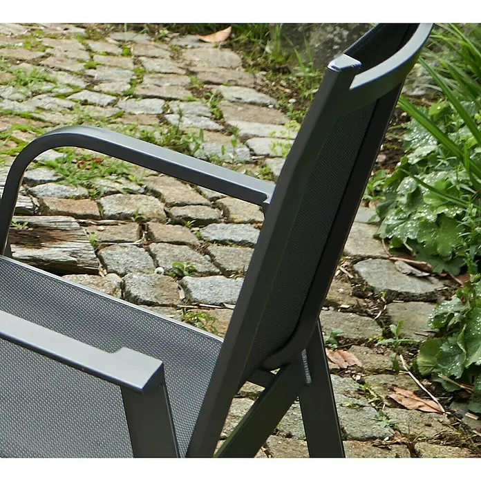 Acamp Stapelsessel Urban - 6 Stk., Aluminium, Acatex, Anthrazit, B x T x H: 62 x 69 x 97 cm - Nahaufnahme eines grauen Gartenstuhls mit Armlehne, der auf einem gepflasterten Weg neben üppigem Grün steht.
