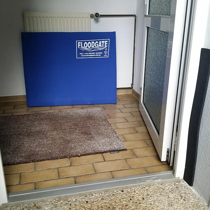 Öko-Tec Dichtungszulage - XL, Selbstklebend, Kürzbar - Blauer Floodgate-Wasserschutz vor einer weißen Wand, ergänzt durch eine Fußmatte und einen teilweise geöffneten Türrahmen.