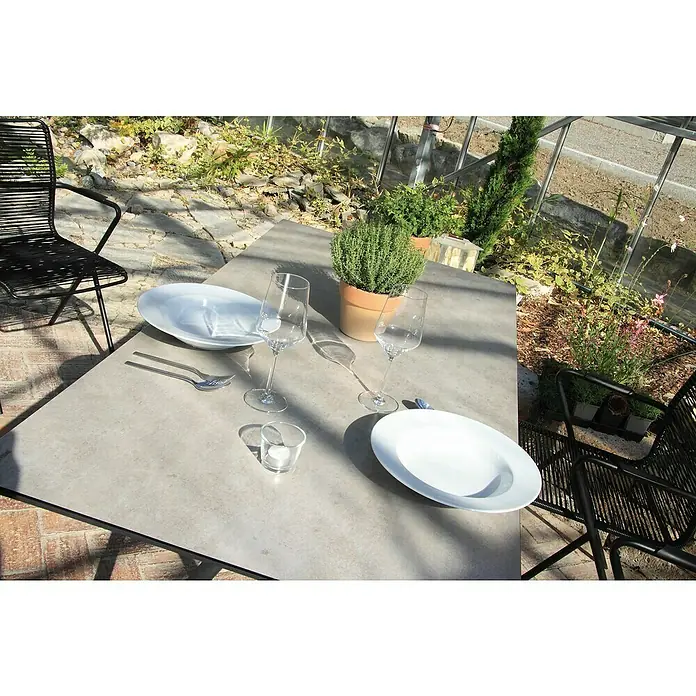 Acamp Gartenmöbel-Set Manhattan/Alpha - 5 -tlg., Aluminium, Glas, Polyester, Stapelbar - Ein gedeckter Tisch mit Tellern, Gläsern und Besteck, dekoriert mit einem Topf mit frischen Kräutern, lädt zum Essen im Freien ein.