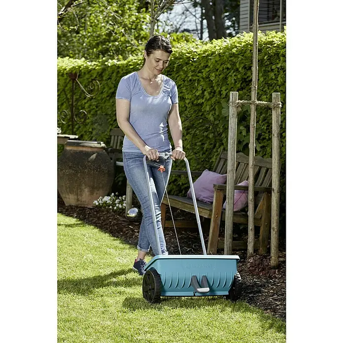 Gardena Streuwagen L - Streubreite: 45 cm, 12,5 l - Eine Frau benutzt einen Gardena Streuwagen im Garten, um Rasen zu säen oder zu düngen. Der Wagen ist blau und hat schwarze Räder.