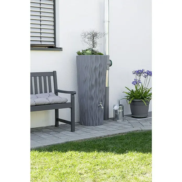 Arves Regenspeicher Alana - 160 l, Betongrau - Graue Regentonne mit Pflanzaufsatz, Wasserhahn und Zubehör, flankiert von Bank und Topfpflanze auf Terrasse mit Rasenfläche.