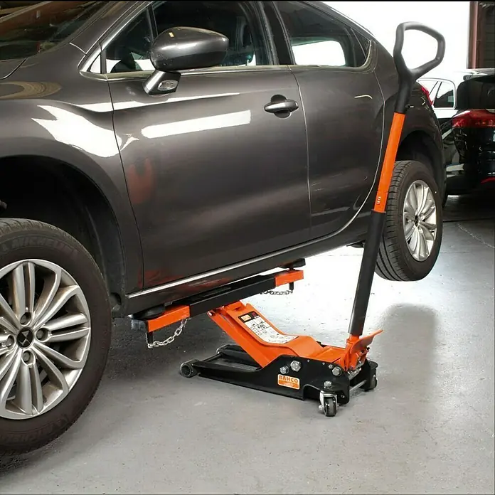 Bahco Hydraulischer Rangierwagenheber BH13000 - Hubhöhe: 90 mm - 550 mm - Das Bild zeigt einen orangefarbenen Bahco Wagenheber, der ein graues Auto in einer Werkstatt anhebt.