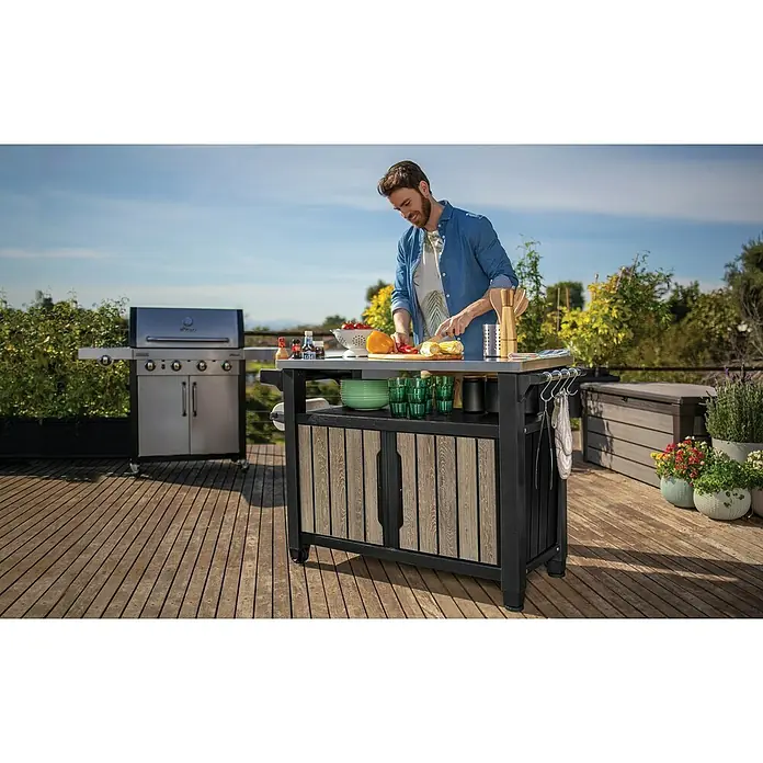Keter Mesa de servicio Unity XL - L x An: 134 x 52 cm, Marrón/Antracita - Un hombre prepara comida en una moderna isla de cocina exterior con una estación de parrilla en un patio de madera.