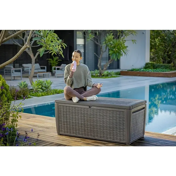 Keter Baúl para jardín Taiwan - L x An x Al: 73 x 145 x 64 cm, Marrón, Plástico - Una mujer sentada en una caja de almacenamiento de plástico con aspecto de ratán Keter. Está bebiendo y relajándose en la piscina.