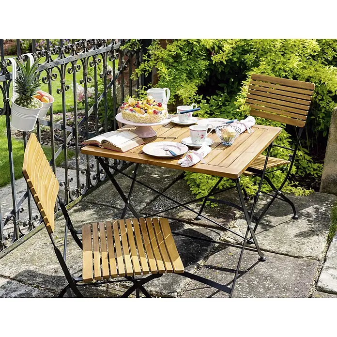 Sunfun Moni Balkontisch - L x B x H: 75 x 75 x 75 cm, Akazie, Klappbar, Eckig, Natur/Schwarz - Gartenszene mit einem Holztisch und zwei Stühlen, gedeckt mit Geschirr, Kuchen und einem Buch, umgeben von Pflanzen im Freien.