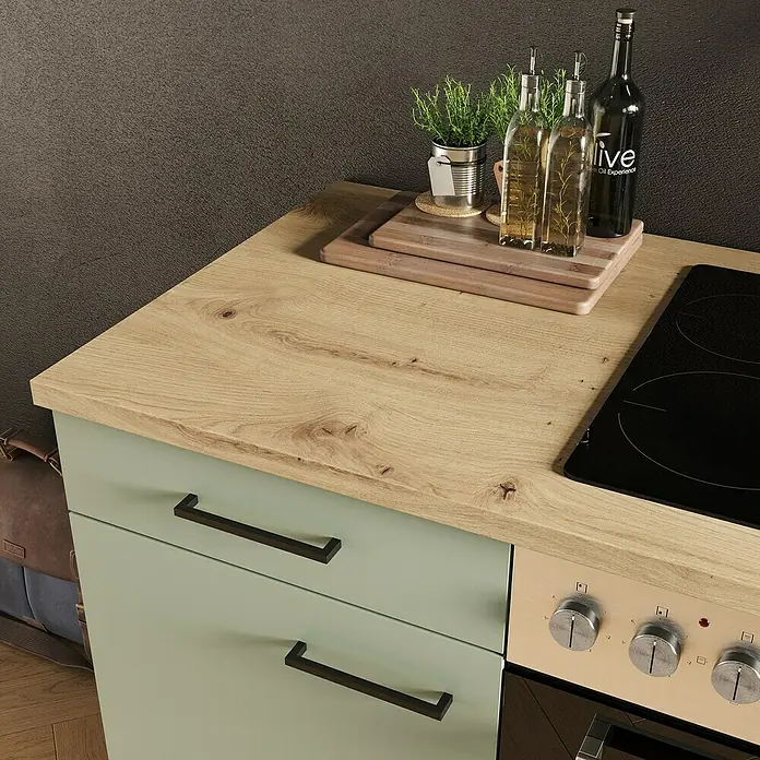 Flex-Well Cara Küchenzeile - Breite: 270 cm, Höhe: 200 cm, Schilfgrün Antifingerprint / Artisan Eiche, Mit Elektrogeräten, Fibonitspüle - Eine Küchenarbeitsplatte aus Holz mit Herd, Gewürzen und Kräutern. Die Schubladen sind in einem hellen Grünton gehalten.