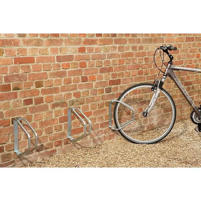 Mottez Aparcamiento de bicicletas - L x An x Al: 285 x 85 x 328 cm, Acero, Montaje en la pared - Un aparcabicicletas de metal con una bicicleta delante, colocado contra una pared de ladrillo sobre grava.