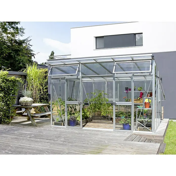 Vitavia Gewächshaus Aura 11500 - Außenmaß inkl. Dachüberstand (B x T): 392,8 x 295 cm, 3 mm, Aluminium - Das Bild zeigt ein Gewächshaus mit geöffneten Fenstern, umgeben von Pflanzen und einer hölzernen Terrasse, vor einem modernen Haus.