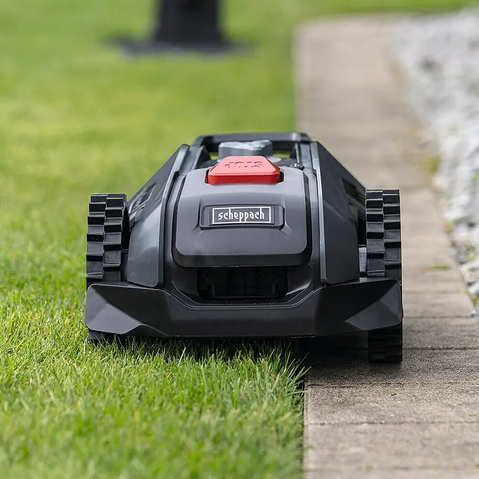 Scheppach Mähroboter RRMA300 - Max. Flächenempfehlung: 300 m², Schnittbreite: 160 mm, Begrenzungskabel - Das Bild zeigt einen Scheppach Rasenmähroboter, der auf einer Rasenfläche neben einem gepflasterten Weg steht.