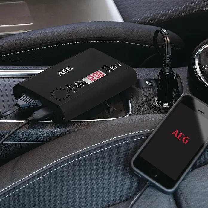 AEG Spannungswandler Si 150 - 230 V, 150 W - Das AEG Ladegerät für das Auto ist abgebildet, mit Kabeln und einem Smartphone, alles im Innenraum eines Autos.