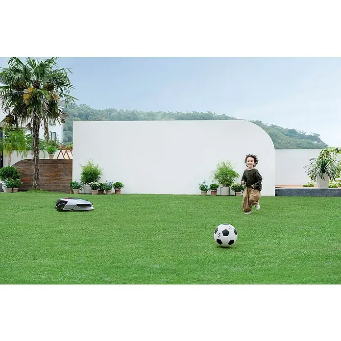 Dreame Mähroboter A2 2000 - 2.000 m², Schnittbreite: 22 cm, Laser-gestützte Navigation, LiDAR - Ein Junge spielt Fußball auf dem Rasen, während ein Rasenmähroboter im Hintergrund arbeitet.
