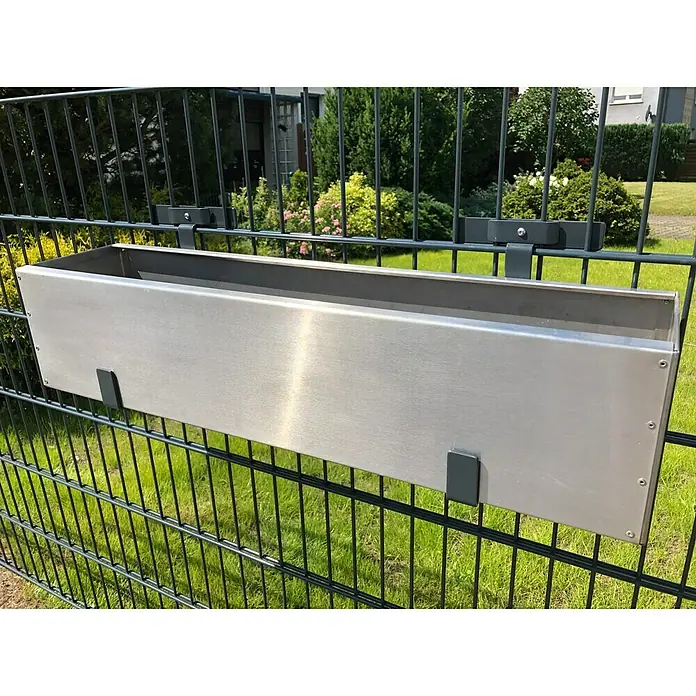 WMT Eindrehhalterung für Blumenkästen - 22 x 17 x 24 cm, Anthrazit - Ein silberner Blumenkasten hängt mit dunklen Halterungen an einem Zaun. Im Hintergrund sind grüne Büsche und ein Rasen zu sehen.