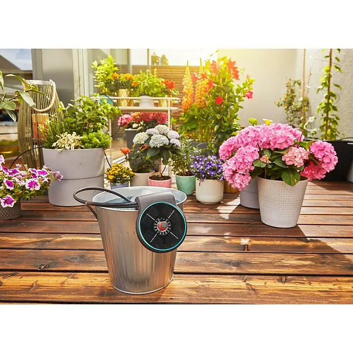 Gardena Bewässerungs-Set AquaBloom - Solarbetrieben - Nahaufnahme eines Gardena Urlaubsbewässerungssystems, das auf einem Eimer sitzt, mit Pflanzen im Hintergrund für automatische Bewässerung.