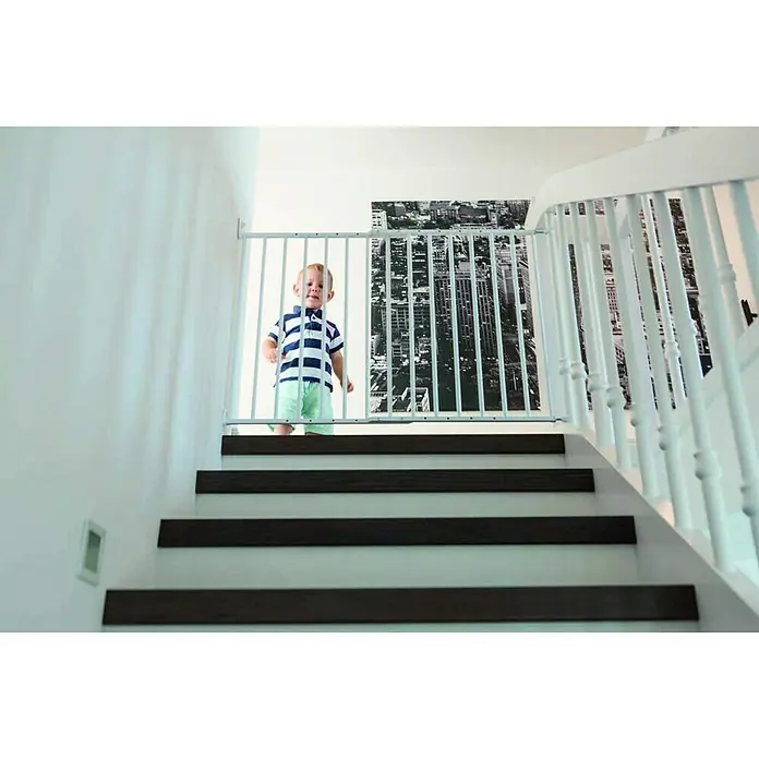 Abus Sicherheitsgitter Tim - Verstellbar: 60 cm - 107 cm - Ein Kleinkind steht hinter einem weißen Treppenschutzgitter, das oben auf einer Treppe mit dunklen Holzstufen angebracht ist.