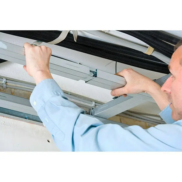Knauf CD-Multiverbinder - 1 Stk. - Ein Mann montiert eine silberne Metallschiene an einer Decke mit Rohren, wodurch ein präzises und professionelles Erscheinungsbild entsteht.