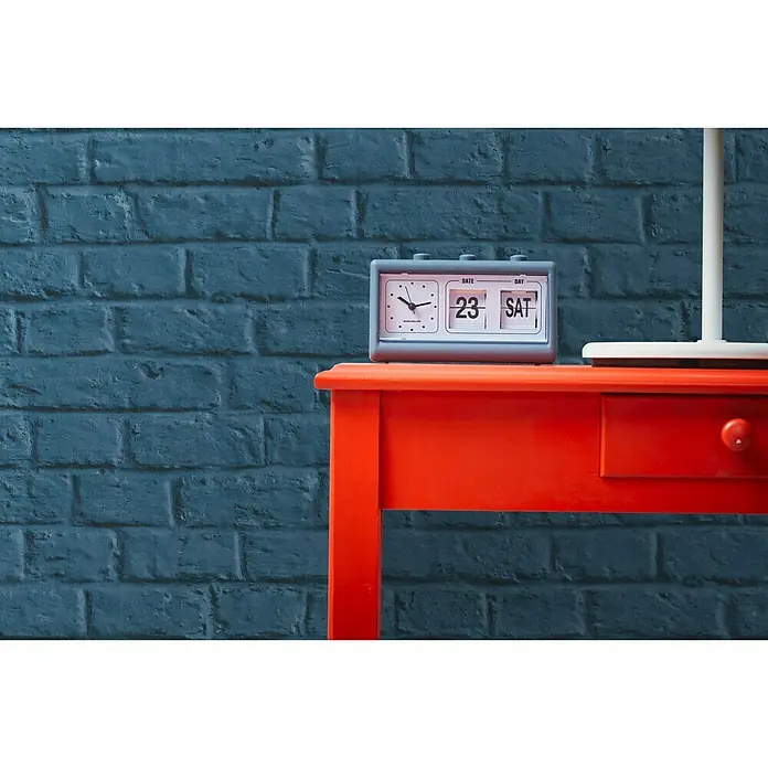 A.S. Création Metropolitan Stories Vliestapete Steine - Blau, Steinoptik, 10,05 x 0,53 m - Ein Vintage-Tischkalender, der auf einem roten Tisch steht, zeigt das Datum "23. Samstag" und Uhrzeit vor einer blauen Ziegelwand.