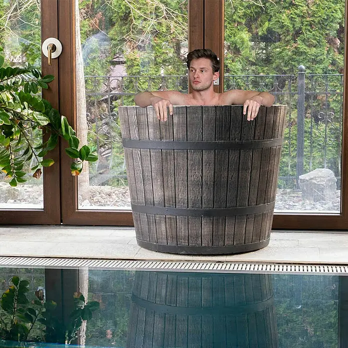 Roto Eistonne Konisch - Ø x H: 900 x 750 cm, Braun, 300 l - Mann entspannt in einer hölzernen Fassbadewanne neben einem Pool, umgeben von üppigem Grün. Die Fassbadewanne ist aus dunklem Holz gefertigt und von Metallreifen umgeben.