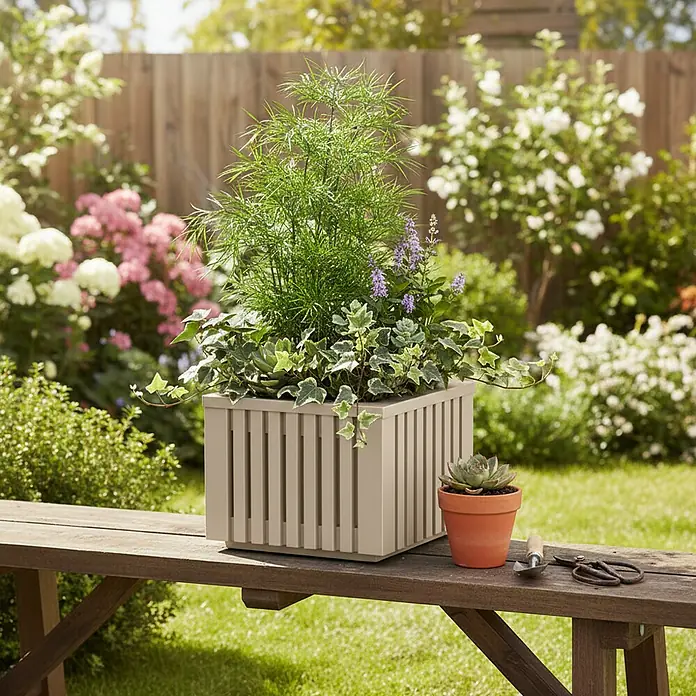 Roto Pflanzkasten Balboa - Kunststoff, Beige - Der quadratische Pflanzkasten aus Holz in Taupe ist mit verschiedenen Pflanzen bepflanzt und dekoriert eine Holzbank im Garten.