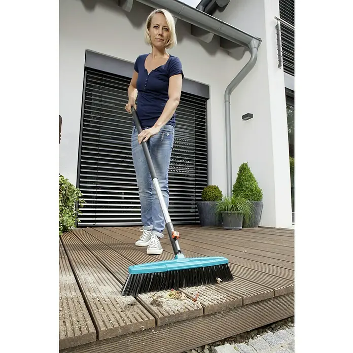 Gardena Combisystem Aluminiumstiel Ergoline 130 - Schwarz/Silber, Material Griff: Kunststoff - Eine Frau benutzt einen GARDENA Besen, um eine Terrasse zu reinigen. Der Besen hat blaue Borsten und einen grauen Stiel.