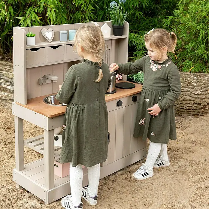 Muddy Buddy Matschküche Explorer - 102 x 40 x 105 cm, Holz, Warmgrau - Zwei Mädchen spielen mit einer Outdoor-Spielküche aus Holz mit Spüle, Herd und Zubehör im Garten.