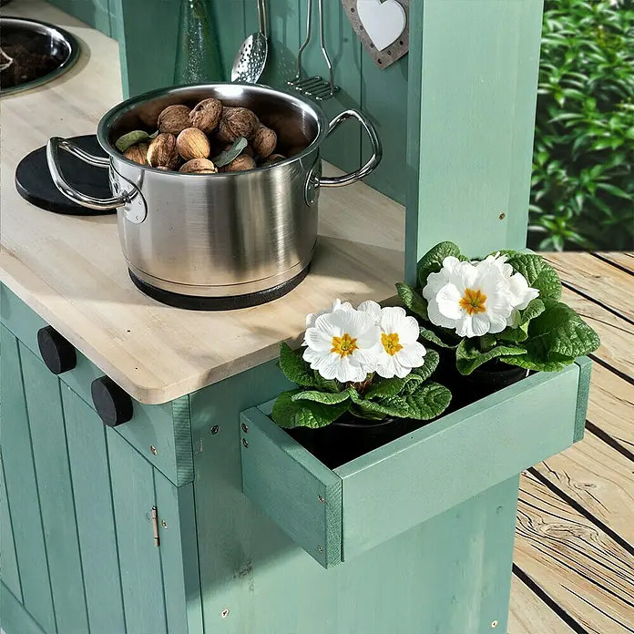 Muddy Buddy Matschküche Explorer - 102 x 40 x 105 cm, Holz, Salbeigrün - Detailaufnahme einer grünen Spielküche mit Topf, Nüssen und Blumenkasten mit weißen Primeln.