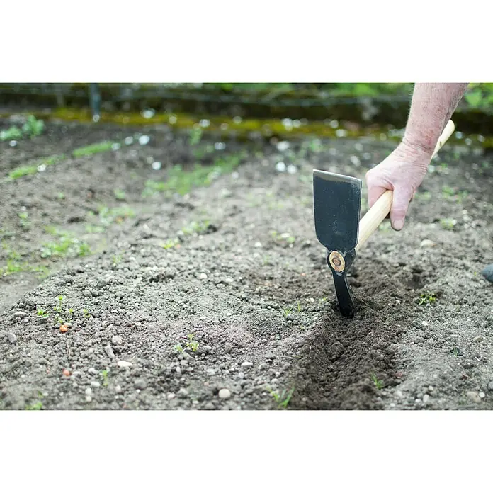 Gardol Kleinhacke - Arbeitsbreite: 7,5 cm, Geschmiedet, Kurzstiel - Eine Hand benutzt eine Gartenhacke mit Holzgriff, um eine Furche in einem Gartenbeet zu ziehen. Die Hacke ist ein nützliches Werkzeug für Gärtner.