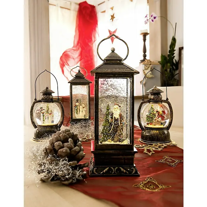 Konstsmide LED-Schneelaterne Schneemannfamilie - Innen, Braun, 12,7 x 10,8 x 20 cm, Schneemann, Batteriebetrieben - Festliche Szene mit Windlichtern mit Schneekugeln, die Weihnachtsmann, Schneemann und Christbaum zeigen, arrangiert auf einem roten Tischläufer mit Tannenzapfen.