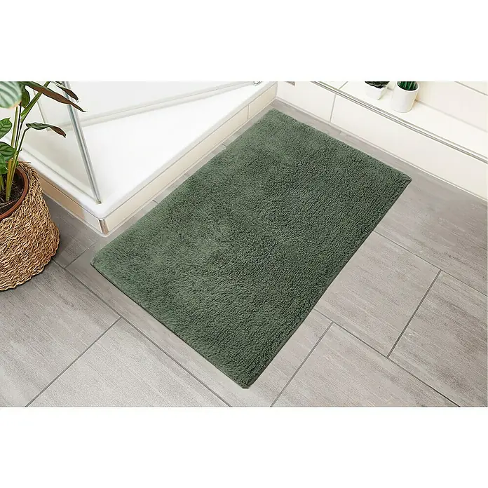 Badteppich Ibiza - 60 x 100 cm, Grün, 100% Baumwolle - Ein rechteckiger, olivgrüner Badteppich liegt auf einem Badezimmerboden mit grauen Fliesen, neben einer Dusche und einer Zimmerpflanze.