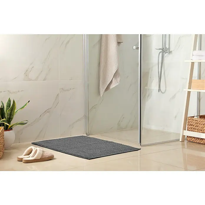 Badteppich Atlanta - 60 x 100 cm, Anthrazit, 100% Baumwolle - Das Badezimmer zeigt eine graue Badematte vor einer Dusche mit Handtuch und Hausschuhen. Ein Regal mit Körben und Handtüchern steht daneben.