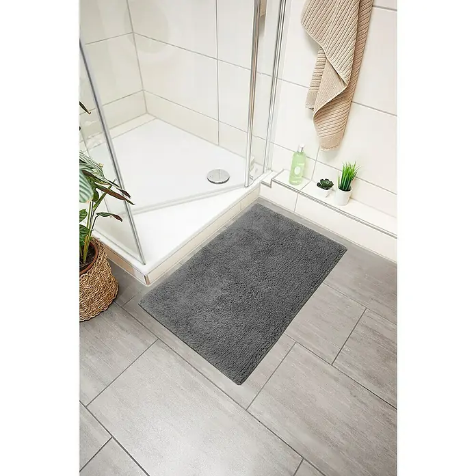 Badteppich Ibiza - 60 x 100 cm, Anthrazit, 100% Baumwolle - Ein grauer, flauschiger Badezimmerteppich liegt vor einer Duschkabine auf hellgrauen Fliesen und sorgt für Komfort und Stil im Badezimmer.